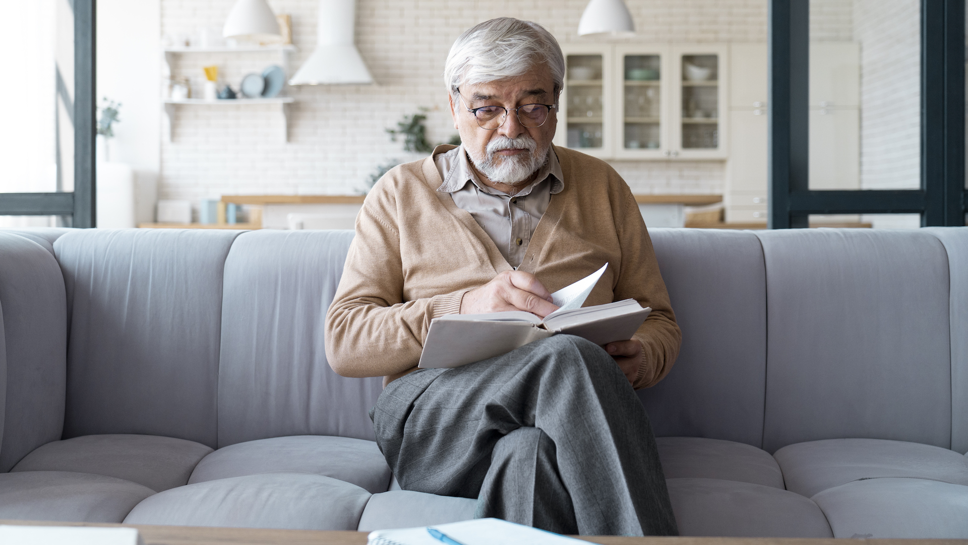 Ein älterer Herr mit grauen Haaren und Brille sitzt konzentriert lesend auf einem Sofa, ein Buch in der Hand haltend, in einem hellen, gemütlich wirkenden Raum.